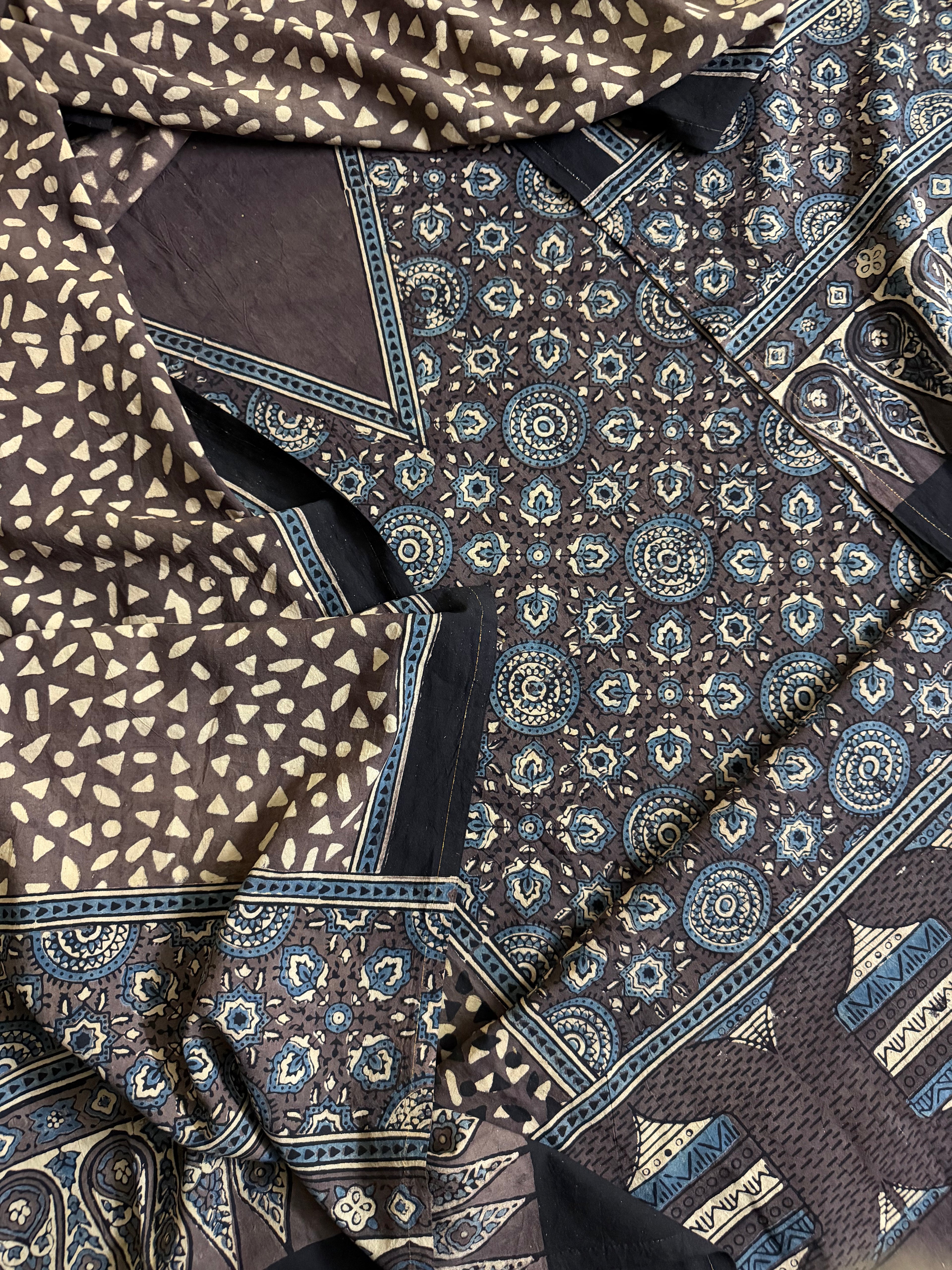 Top-down flat lay of brown Ajrakh block printed cotton kurta fabric with indigo medallion and floral motifs, part of 3-piece unstitched suit set