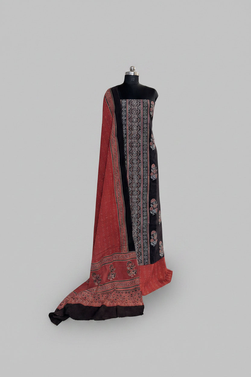 Black and red Ajrakh cotton 3-piece unstitched suit set on mannequin — deep black kurta with geometric block print border, red dupatta with floral Ajrakh print and tassel hem, and black bottom fabric