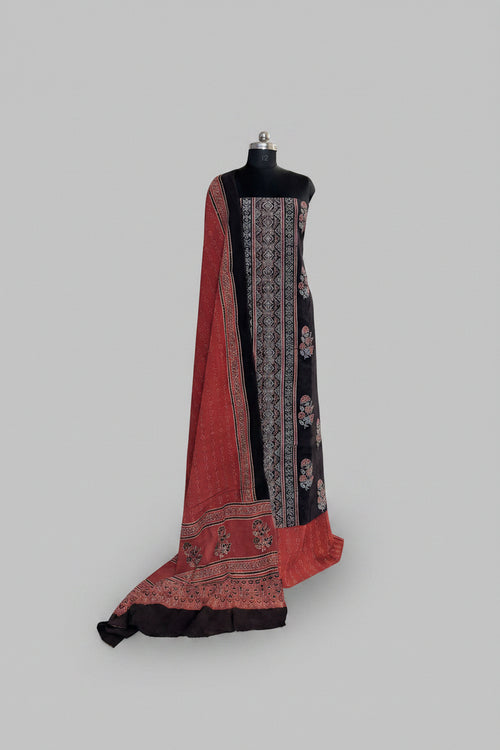 Black and red Ajrakh cotton 3-piece unstitched suit set on mannequin — deep black kurta with geometric block print border, red dupatta with floral Ajrakh print and tassel hem, and black bottom fabric
