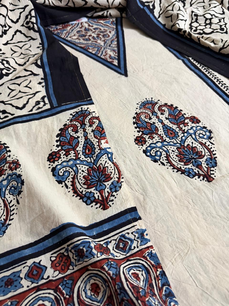 Close-up of oatmeal Ajrakh cotton kurta V-neckline with blue floral inset, large blue and red paisley medallion body print, and navy blue border stripe with geometric Ajrakh dupatta in background