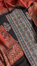 Flat lay close-up of black and red Ajrakh hand block printed cotton 3-piece unstitched suit set showing black kurta with geometric diamond border, floral motifs, red dupatta with paisley print, and black salwar