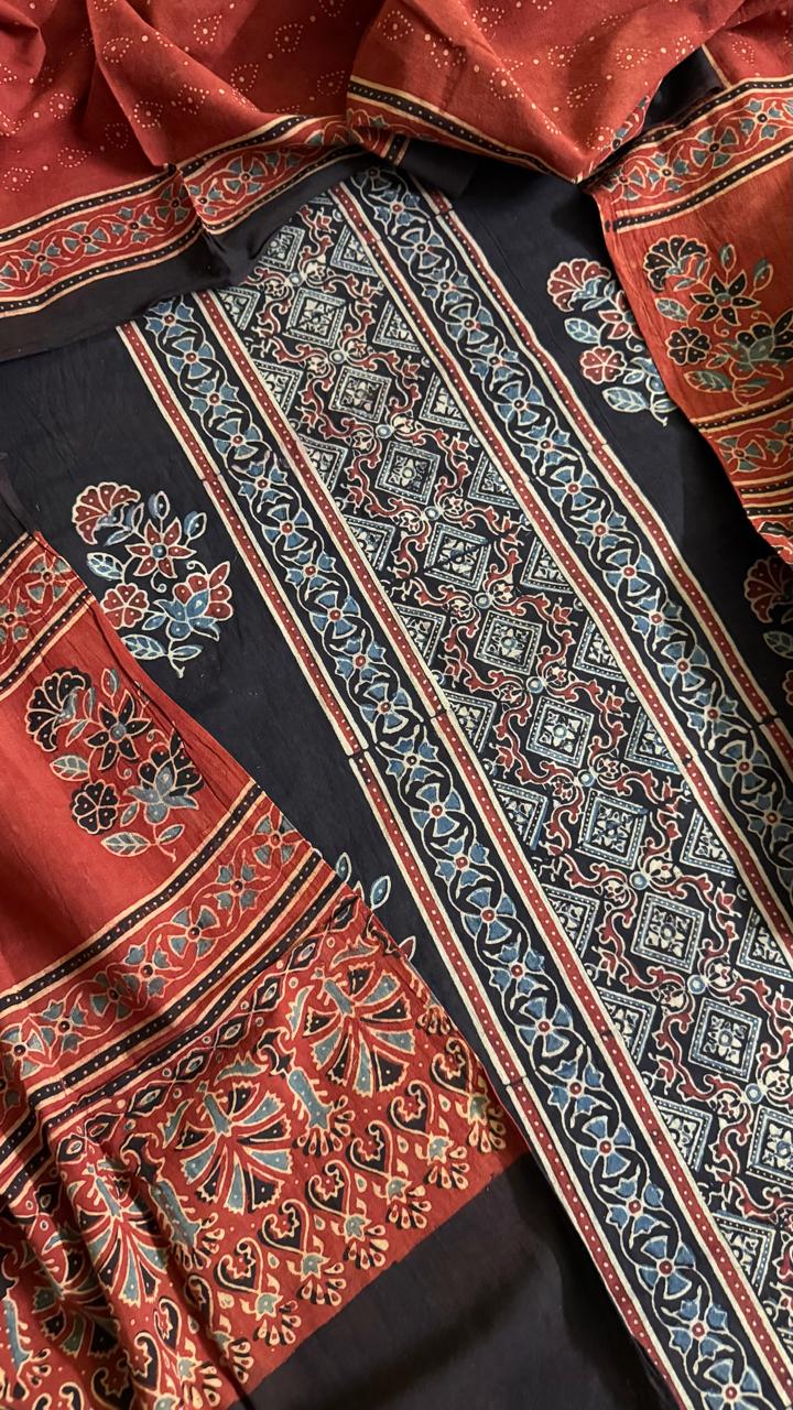 Flat lay close-up of black and red Ajrakh hand block printed cotton 3-piece unstitched suit set showing black kurta with geometric diamond border, floral motifs, red dupatta with paisley print, and black salwar