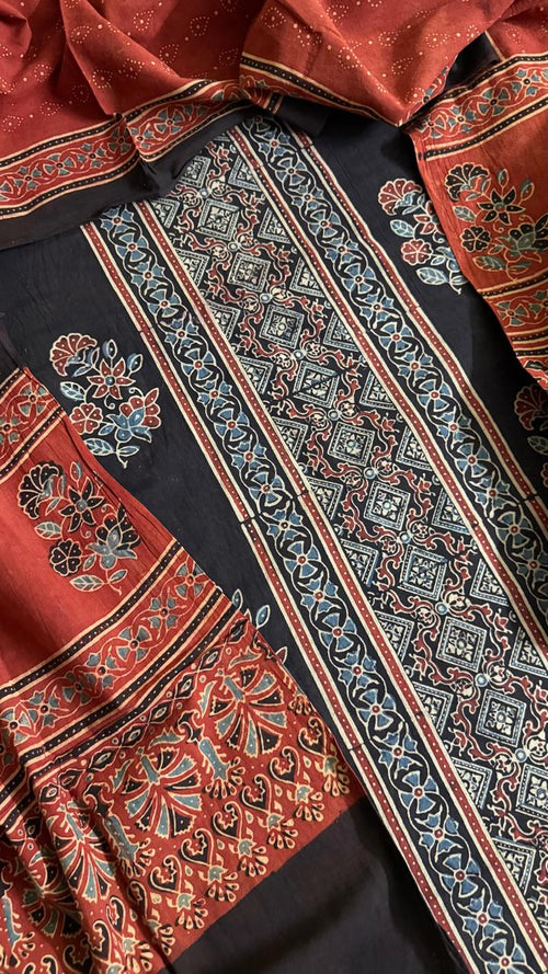 Flat lay close-up of black and red Ajrakh hand block printed cotton 3-piece unstitched suit set showing black kurta with geometric diamond border, floral motifs, red dupatta with paisley print, and black salwar