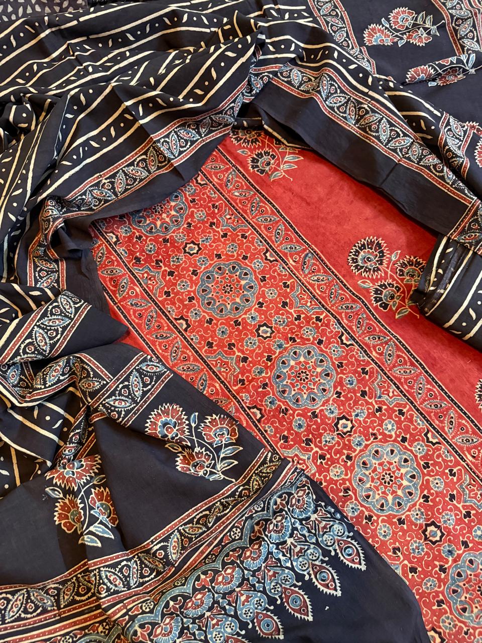 Close-up flat lay of red and black Ajrakh cotton 3-piece unstitched suit set — red kurta with dense medallion and floral block print, black dupatta with diagonal stripe and paisley border, and plain red bottom fabric