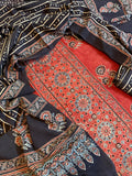 Close-up flat lay of red and black Ajrakh cotton 3-piece unstitched suit set — red kurta with dense medallion and floral block print, black dupatta with diagonal stripe and paisley border, and plain red bottom fabric