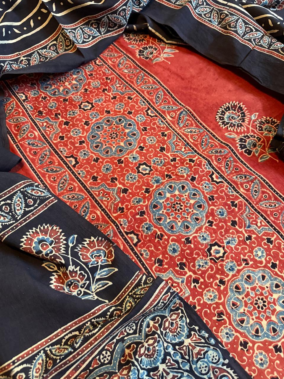 Close-up of deep red Ajrakh cotton kurta fabric with large blue and black medallion hand block print, dense floral fill motifs, and black border with carnation flower print