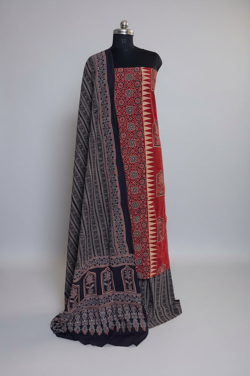 Red and black Ajrakh cotton 3-piece unstitched suit set on mannequin — deep red kurta with geometric star lattice block print and zigzag border, draped with black Ajrakh dupatta featuring floral arch border