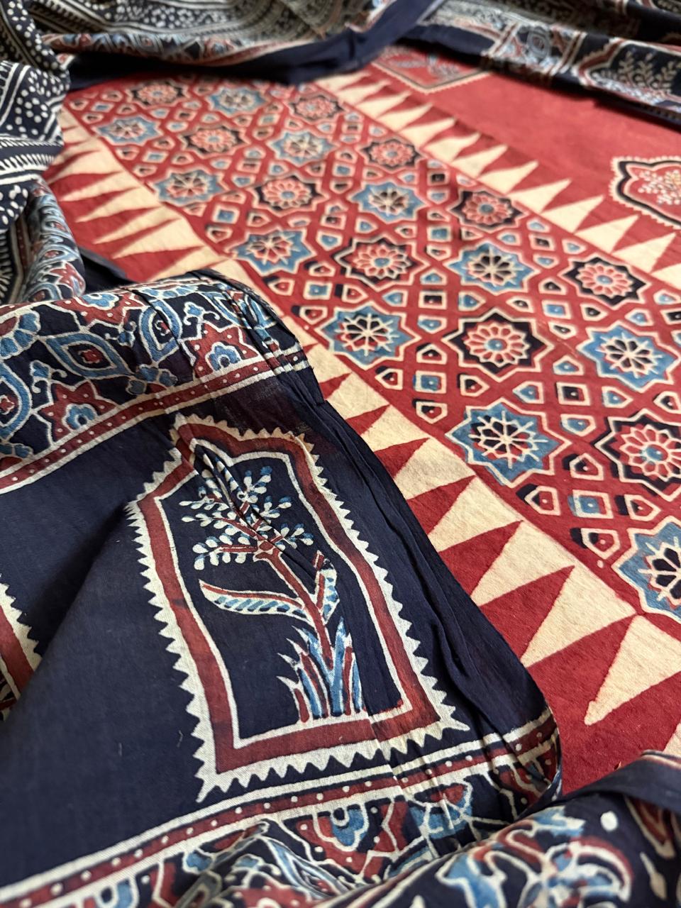 Close-up of black Ajrakh dupatta with large floral arch motif block print alongside red cotton kurta fabric with geometric star lattice and cream zigzag border"
