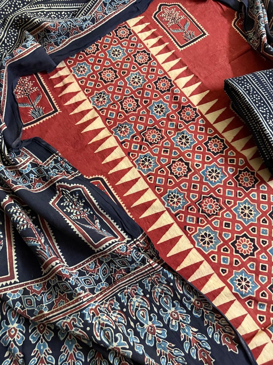 Flat lay of red Ajrakh cotton kurta fabric with geometric star lattice and zigzag border block print, floral arch panels, and black Ajrakh dupatta with paisley and geometric fill motifs