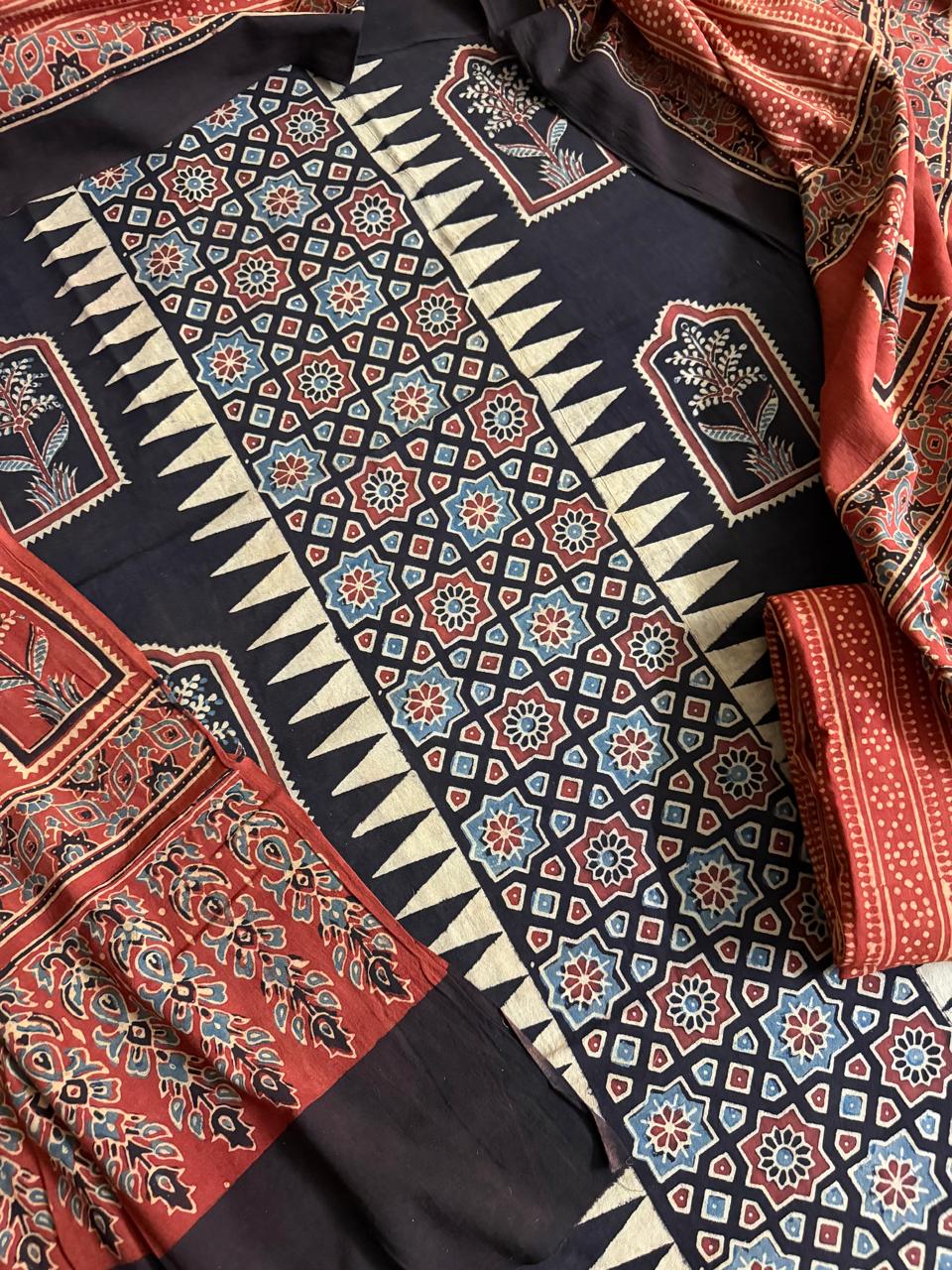 Top-down flat lay of black Ajrakh cotton kurta with geometric star lattice and zigzag border hand block print, floral arch panels, and red Ajrakh dupatta with dense floral motif scatter