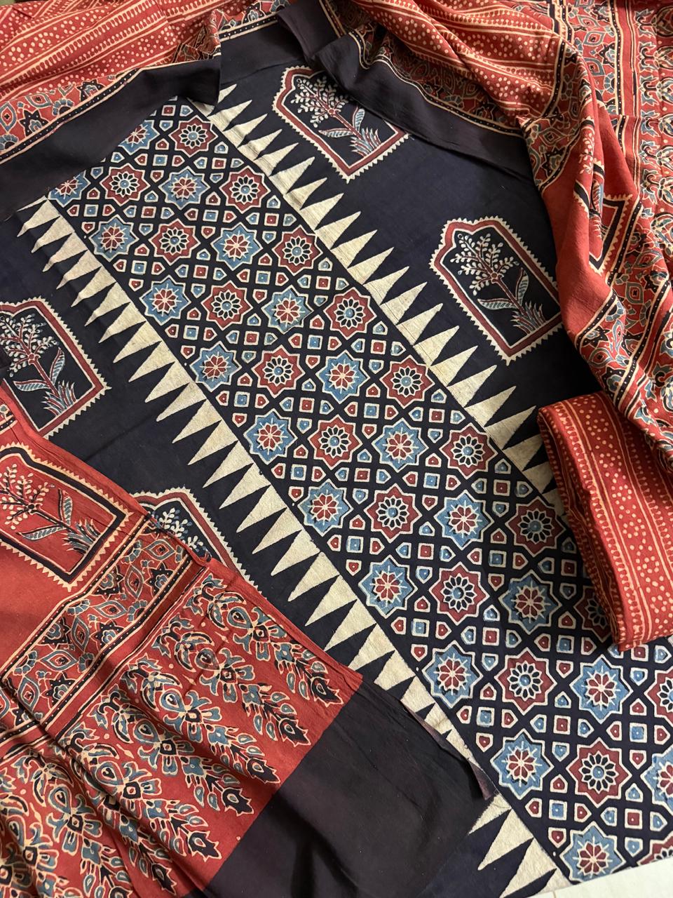 Flat lay of black Ajrakh cotton kurta fabric with geometric star lattice and cream zigzag border block print, floral arch motif panels, alongside red Ajrakh dupatta with dense floral and paisley print