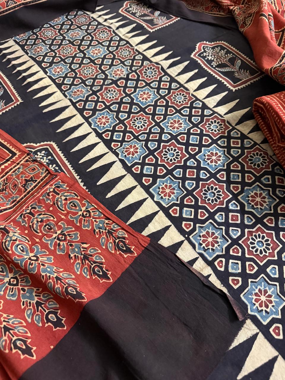 Close-up of black Ajrakh cotton kurta with geometric star lattice block print and cream zigzag border, with red Ajrakh dupatta featuring large floral and leaf motifs in blue and green tones
