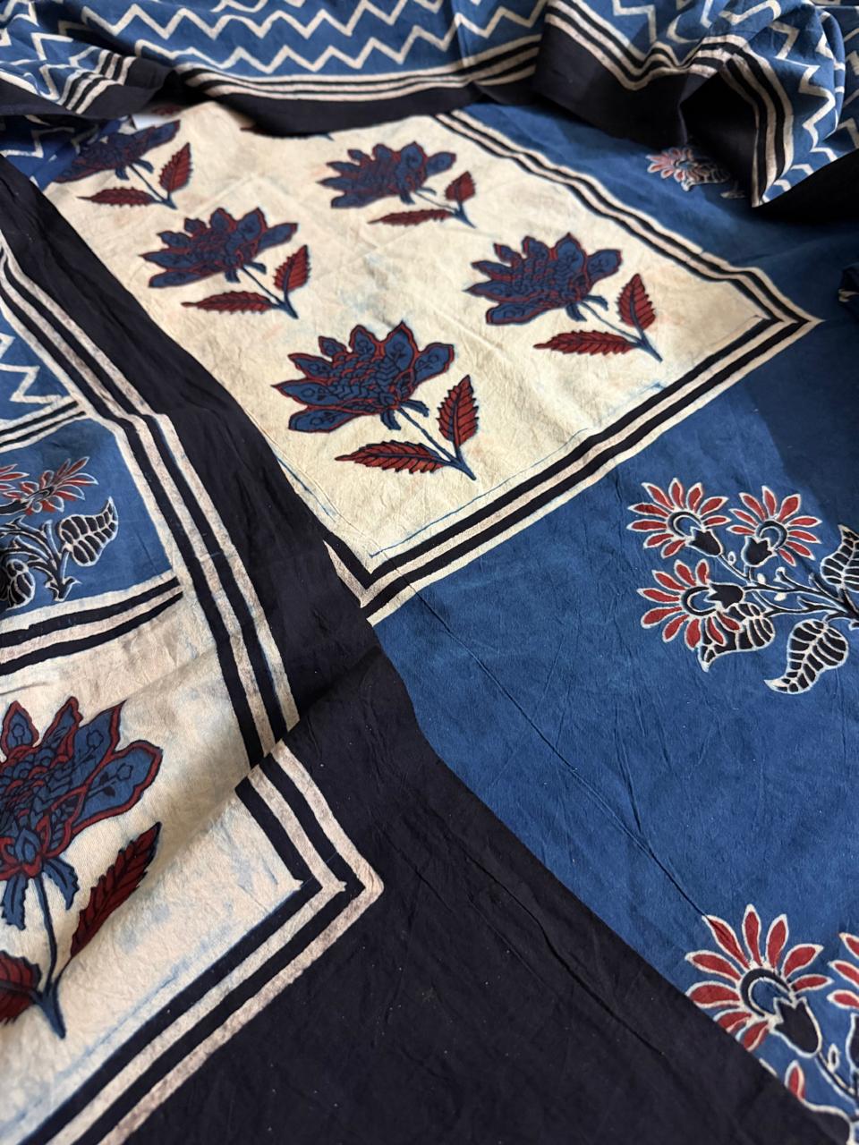 Close-up of black Ajrakh cotton kurta with cream yoke panel showing blue and red lotus floral block print, indigo blue body with scattered sunflower and floral motifs, and blue zigzag chevron dupatta border