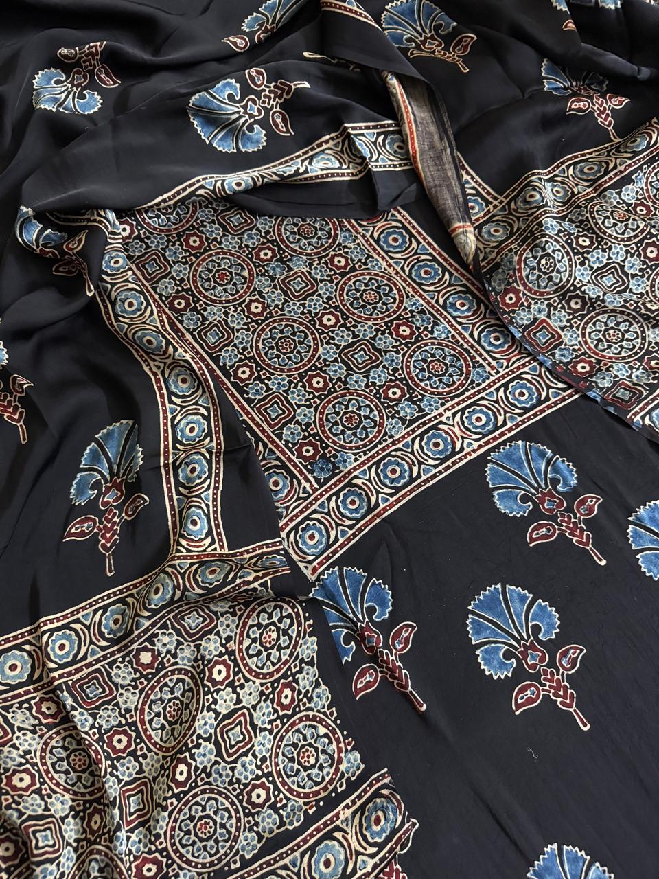 Close-up flat lay of black Ajrakh Modal silk fabric showing blue and maroon geometric circle and diamond medallion yoke, blue glory lily floral body print
