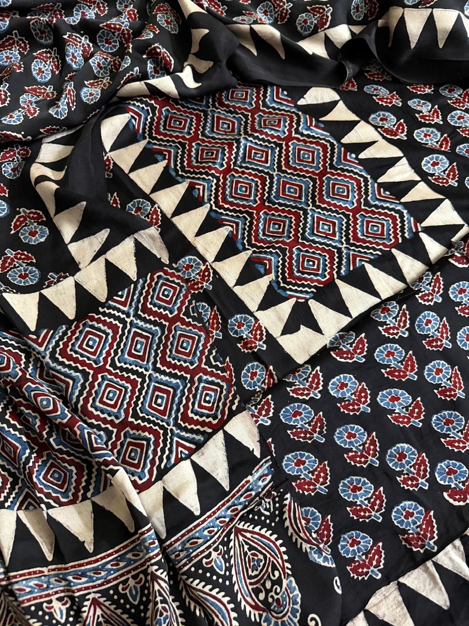Close-up of black Ajrakh modal silk fabric pieces showing red and blue geometric diamond yoke, floral body print and paisley dupatta border
