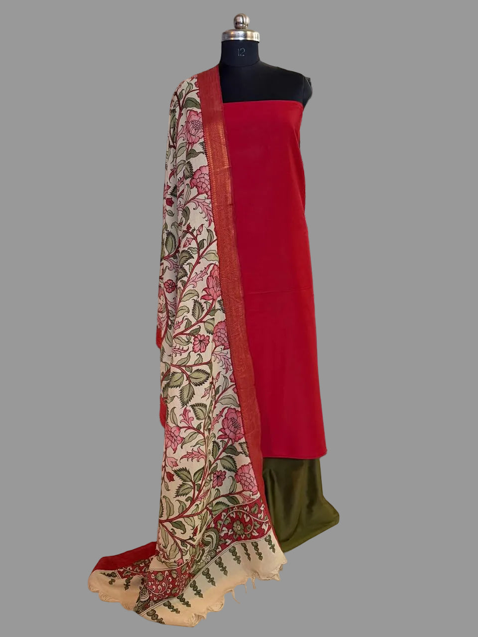 Mannequin display of 3-piece unstitched suit set – red cotton silk kurta with hand pen Kalamkari ivory dupatta draped over shoulder showing floral body and peacock pallu, forest green cotton silk bottom