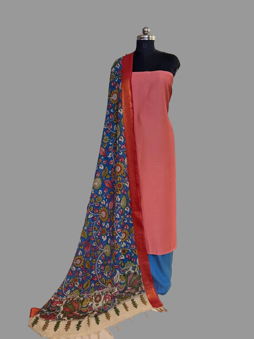 Mannequin display of 3-piece unstitched suit set – rose pink cotton silk kurta with hand painted blue Kalamkari dupatta draped over shoulder, showing multicolour floral body and ivory peacock pallu border