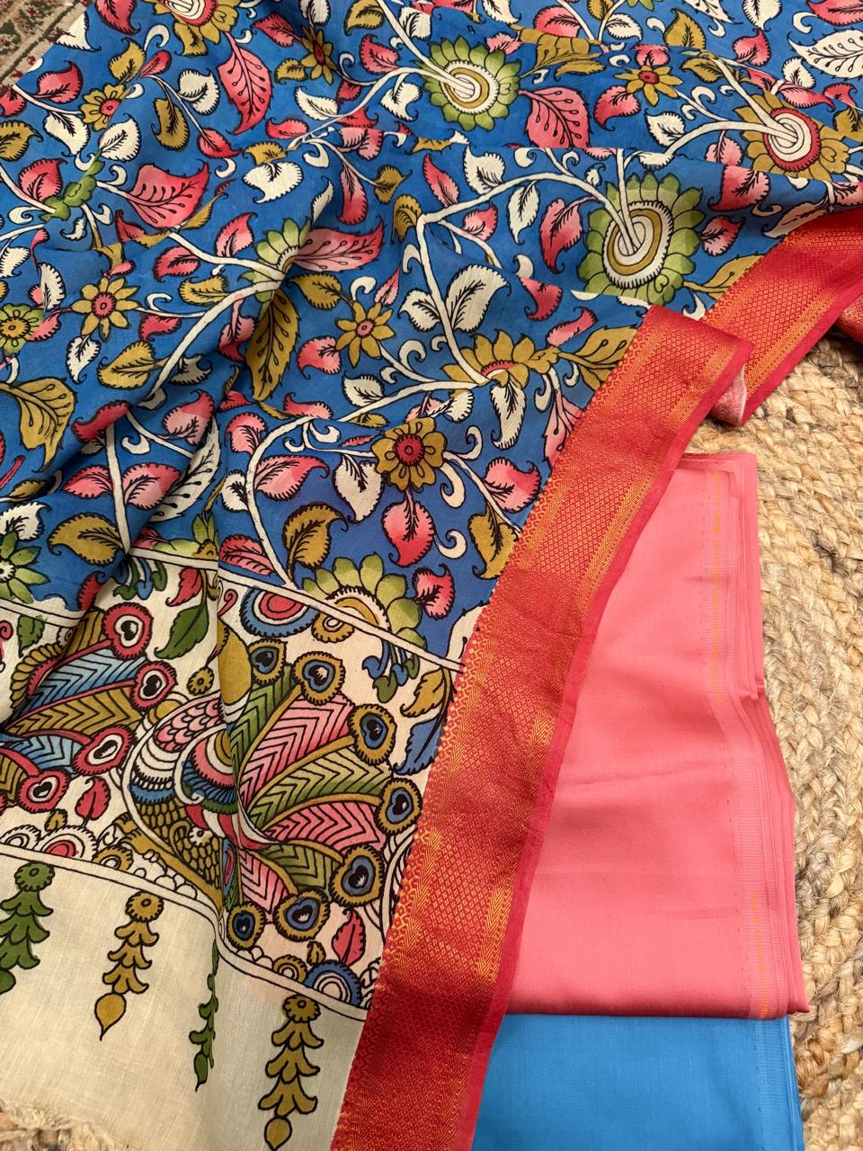 Overhead flat lay of folded rose pink cotton silk and slate blue cotton silk suit fabrics framed by hand painted blue Kalamkari dupatta – ivory peacock pallu and red-gold Zari border visible at edge