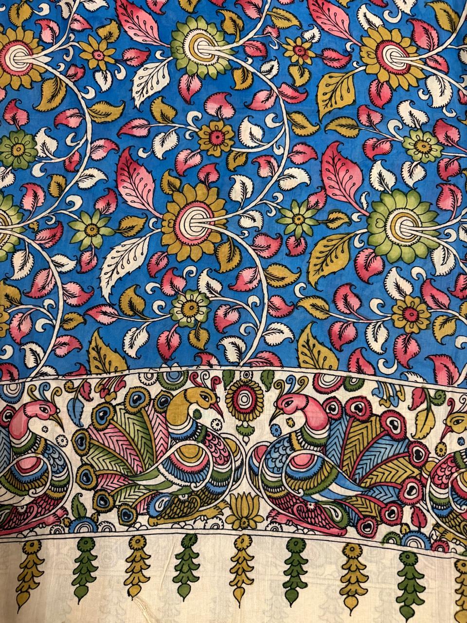 Close-up of hand painted Kalamkari dupatta body-to-border transition – dense cobalt blue ground with pink lotus, gold sunflower, and white foliage vine above ivory peacock-pair border with tassel fringe