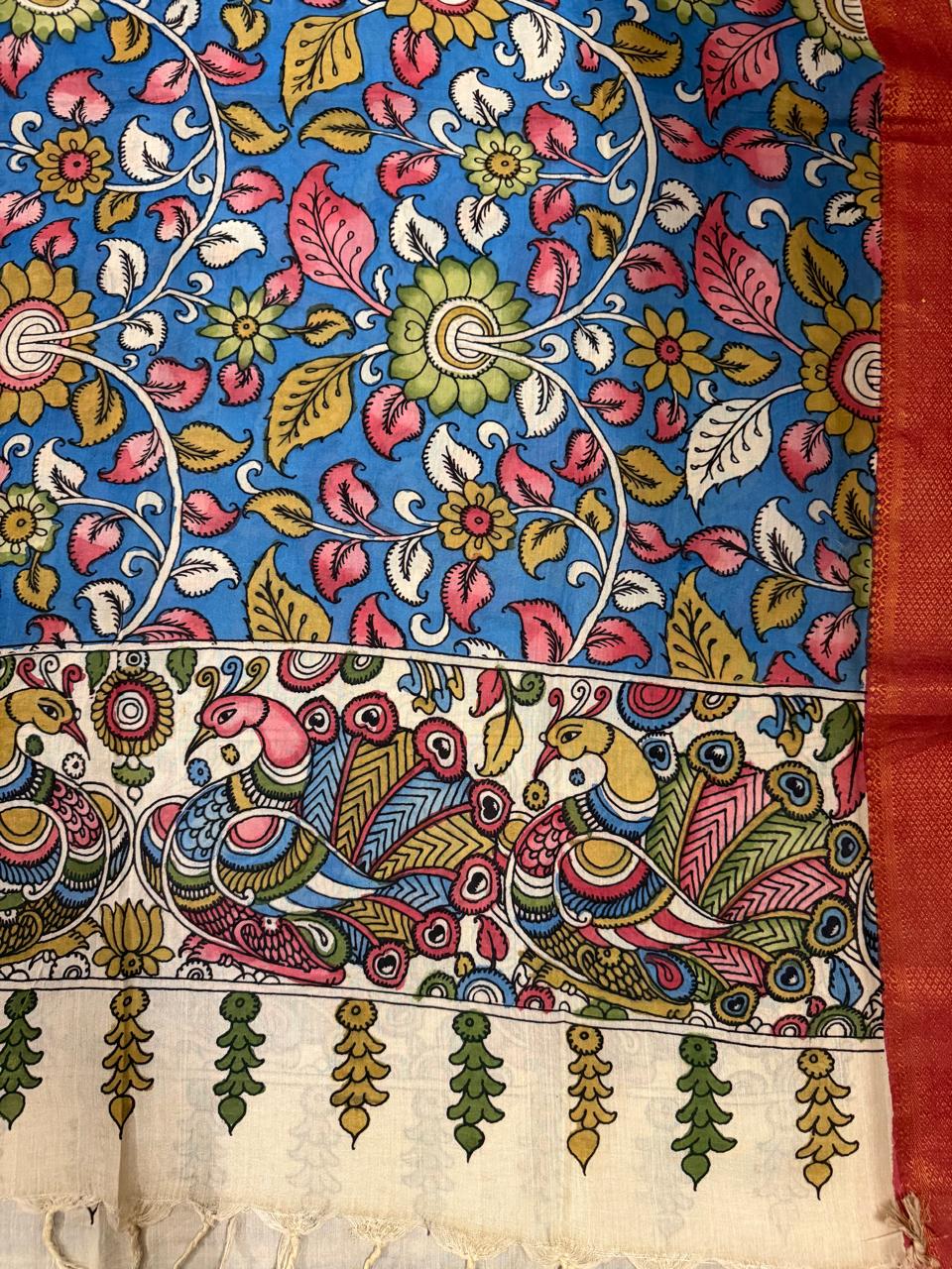 Flat lay detail of hand painted Kalamkari dupatta – cobalt blue ground dense pink lotus, gold sunflower, and white scroll floral vine body transitioning to ivory peacock-pair border with red-gold Zari edge and tassel fringe