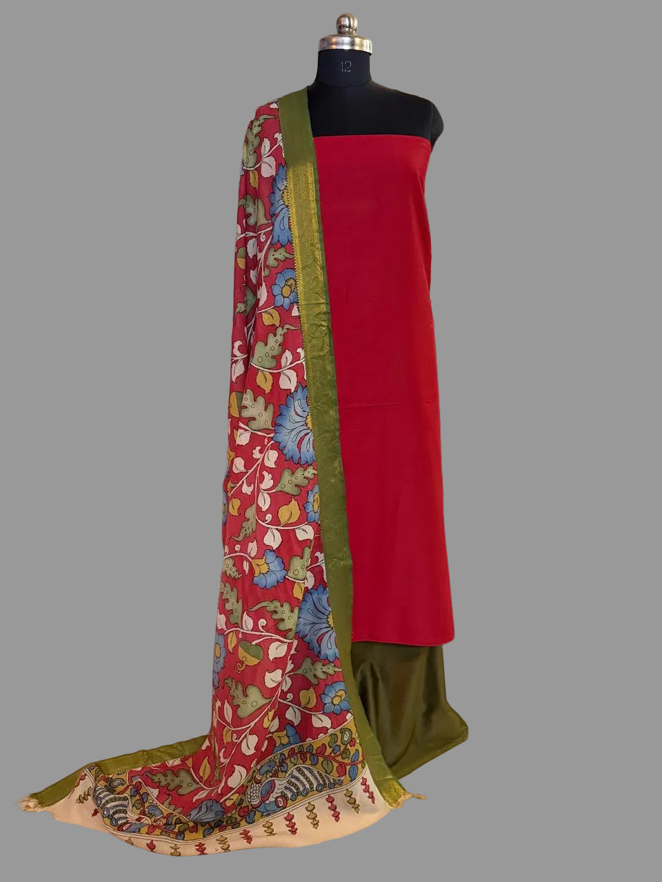 Mannequin display of 3-piece unstitched suit set – red cotton silk kurta with hand pen Kalamkari crimson dupatta draped over shoulder, green gold Zari border visible, forest green cotton silk bottom