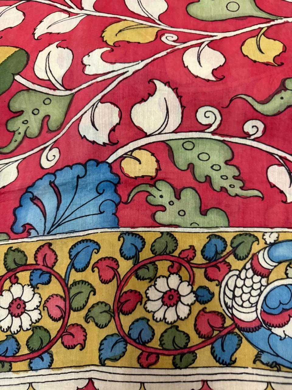 Macro detail of hand pen Kalamkari dupatta border transition – crimson red body with blue leaf and white floral vine above yellow-gold peacock-motif border with pink and white floral scroll, Mangalgiri silk