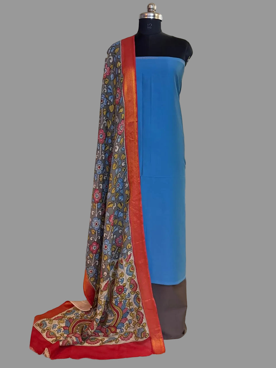 Mannequin display of 3-piece unstitched suit set – state blue cotton silk kurta with hand painted grey Kalamkari dupatta draped over shoulder showing floral body and red-orange Zari border, charcoal brown bottom