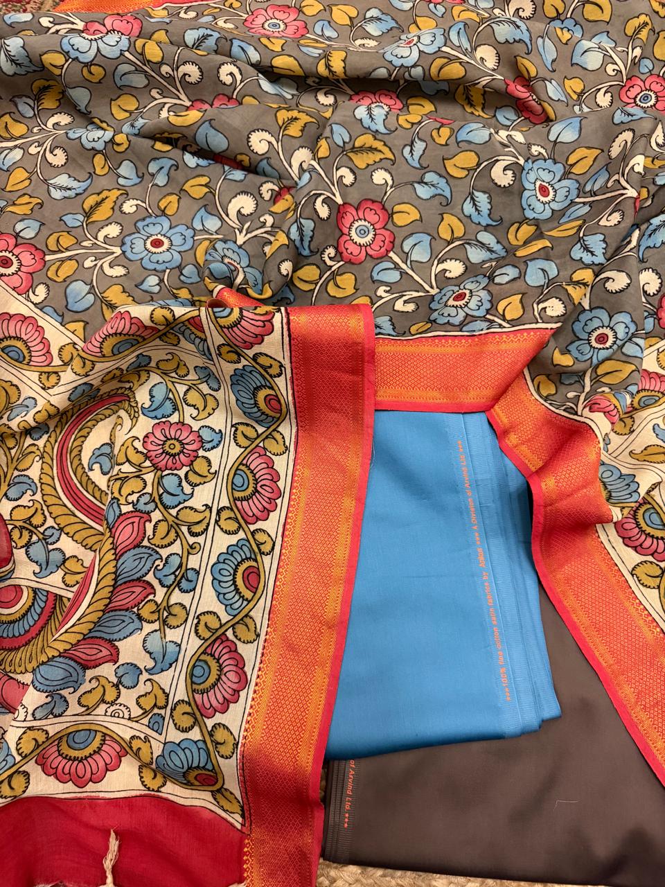 Flat lay detail showing red-orange Zari border of hand painted Kalamkari dupatta alongside folded state blue cotton silk kurta and charcoal brown bottom – grey floral body spread behind, House of Tantuvana