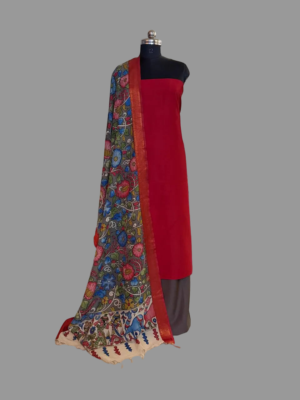 Mannequin display of 3-piece unstitched suit set – vibrant red cotton silk kurta with hand painted grey Kalamkari dupatta draped over shoulder showing dense floral body and red-gold Zari border, charcoal bottom