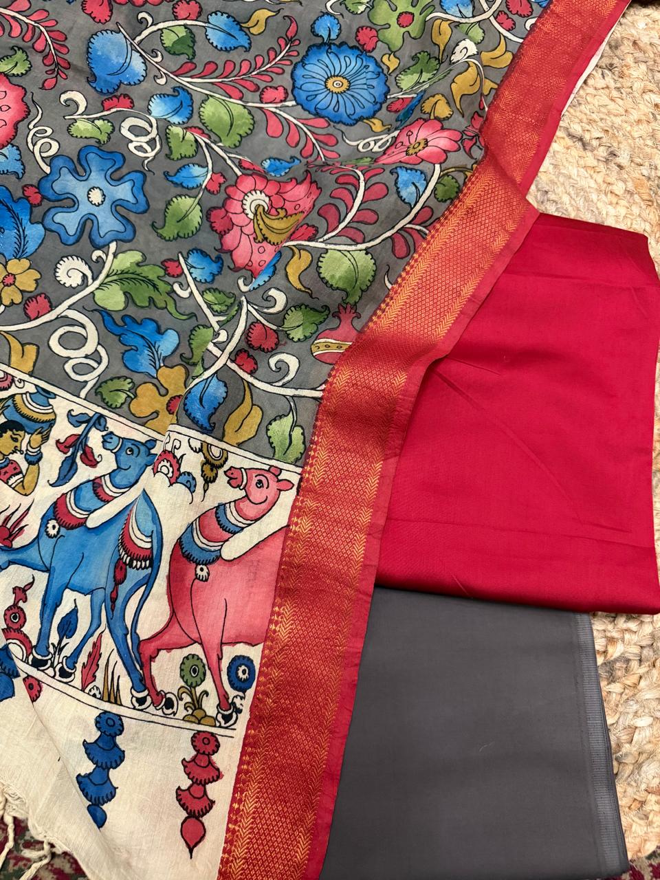 Flat lay showing hand painted Kalamkari dupatta grey body and ivory mythological border alongside red cotton silk and charcoal grey bottom – blue camel and pink animal figures on ivory pallu, red-gold Zari border