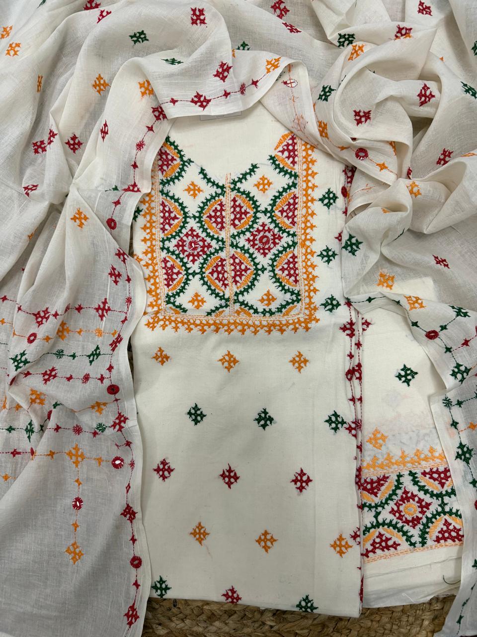 Angled flatlay of cream cotton kurta fabric showing multicolour Sindhi hand-embroidered neckline panel and hemline border in green, red and mustard threads