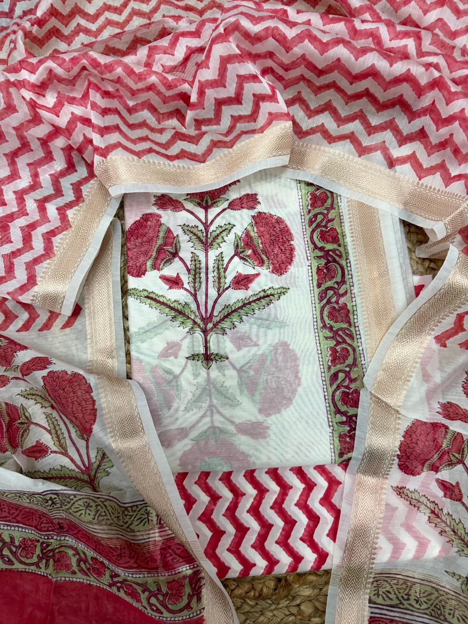Straight overhead flatlay of white cotton Mughal block print unstitched suit set – red marigold floral kurta panel with gold zari side border, red chevron zigzag bottom and dupatta with floral border