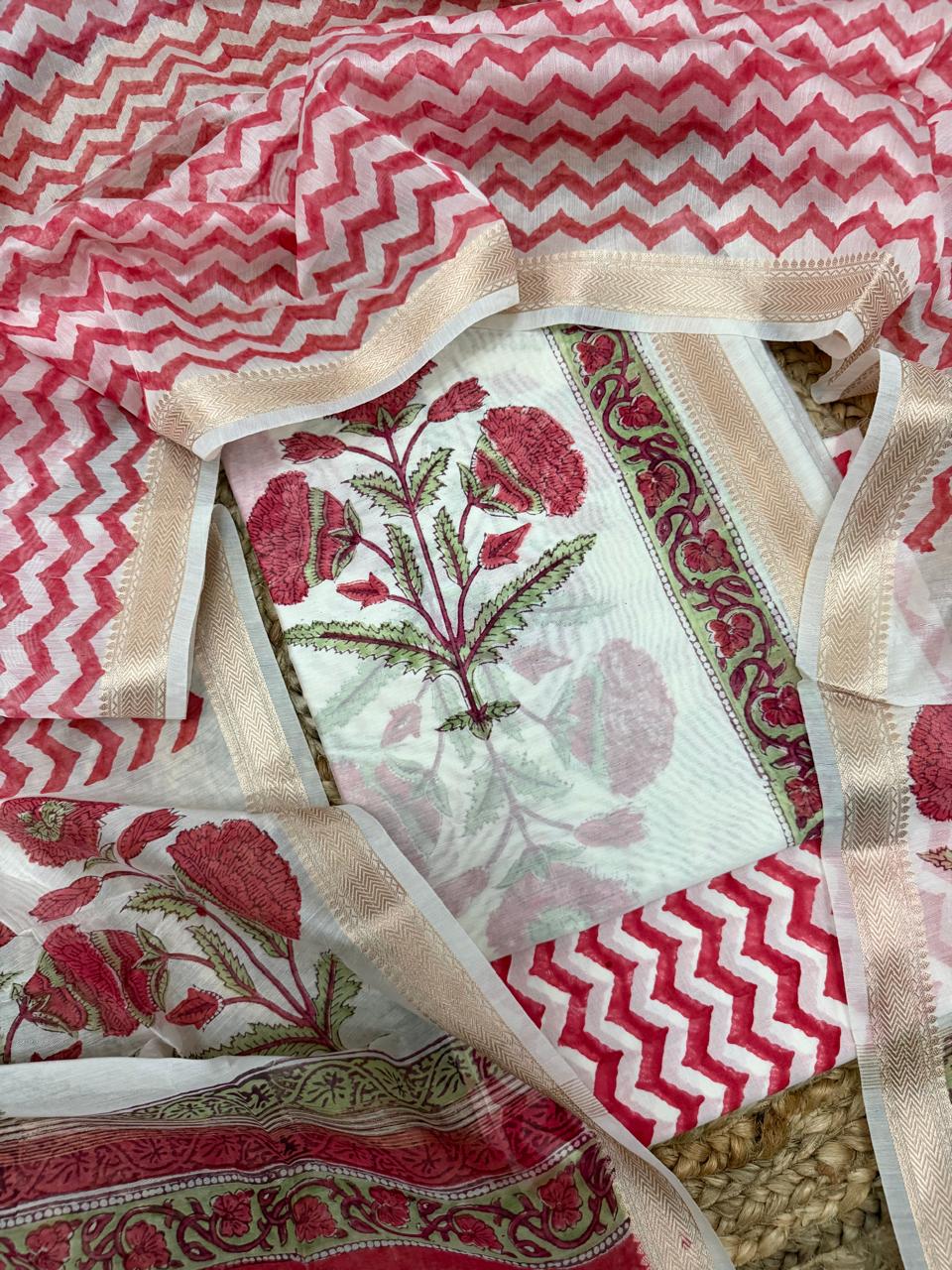 Angled overhead flatlay of white cotton Mughal block print unstitched suit set – red marigold floral kurta panel with gold zari border, red chevron bottom and dupatta, 3-piece suit set