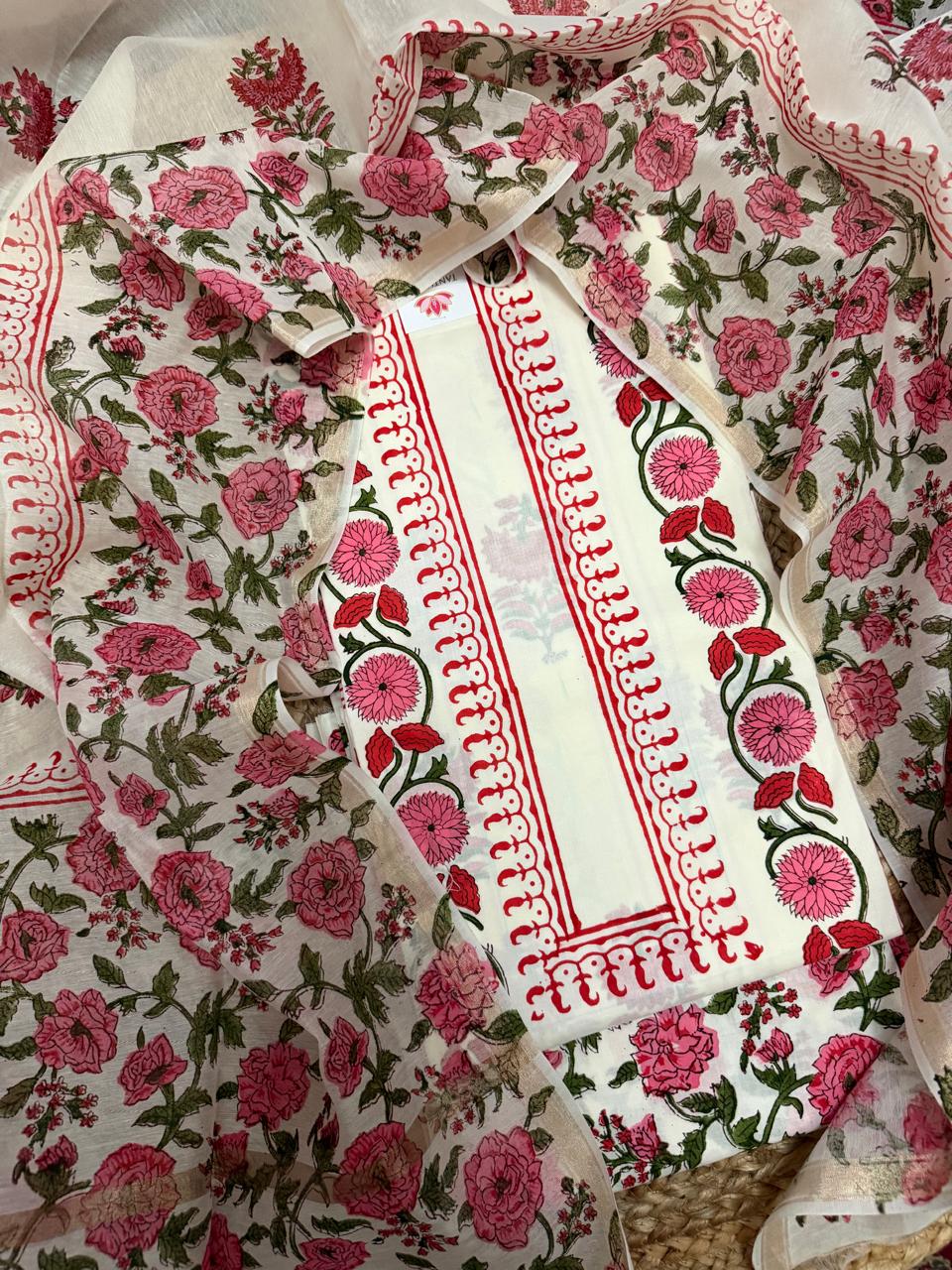 Angled overhead flatlay of white cotton placement floral block print unstitched kurta – pink dahlia and red tulip vine border strip with red scallop edge, Chanderi silk dupatta with pink rose and green leaf all-over print draped 