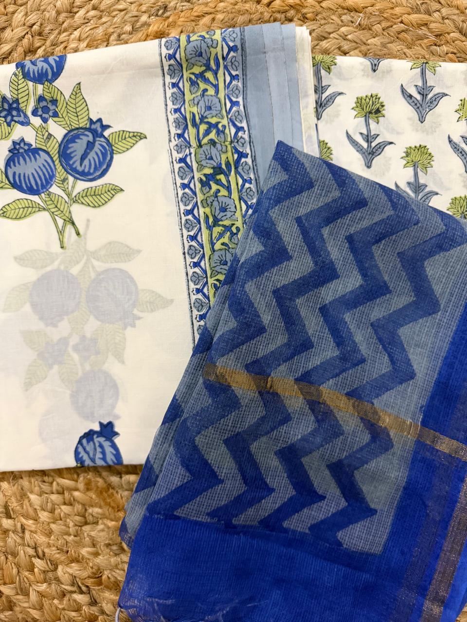 Angled flatlay of white cotton pomegranate block print kurta fabric with blue floral border strip, paired with blue chevron zigzag Kota Doria dupatta with gold zari, unstitched 3-piece suit set