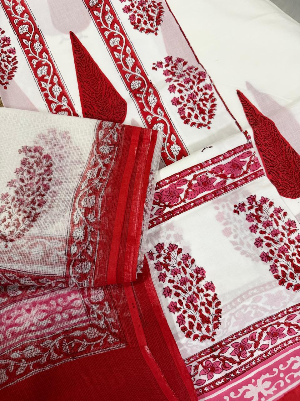 Angled flatlay of white cotton placement block print 3-piece unstitched suit set – red paisley teardrop kurta with floral vine border strips, pink floral bottom fabric and sheer Kota Doria dupatta with red floral vine border folded in foreground