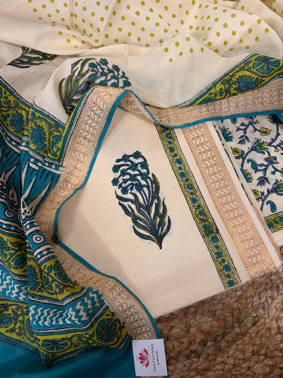 Close flat lay of cream cotton block print kurta fabric with teal-green floral bouquet block print motif, golden border with teal piping, alongside teal block print dupatta and cream polka dot fabric on jute mat 