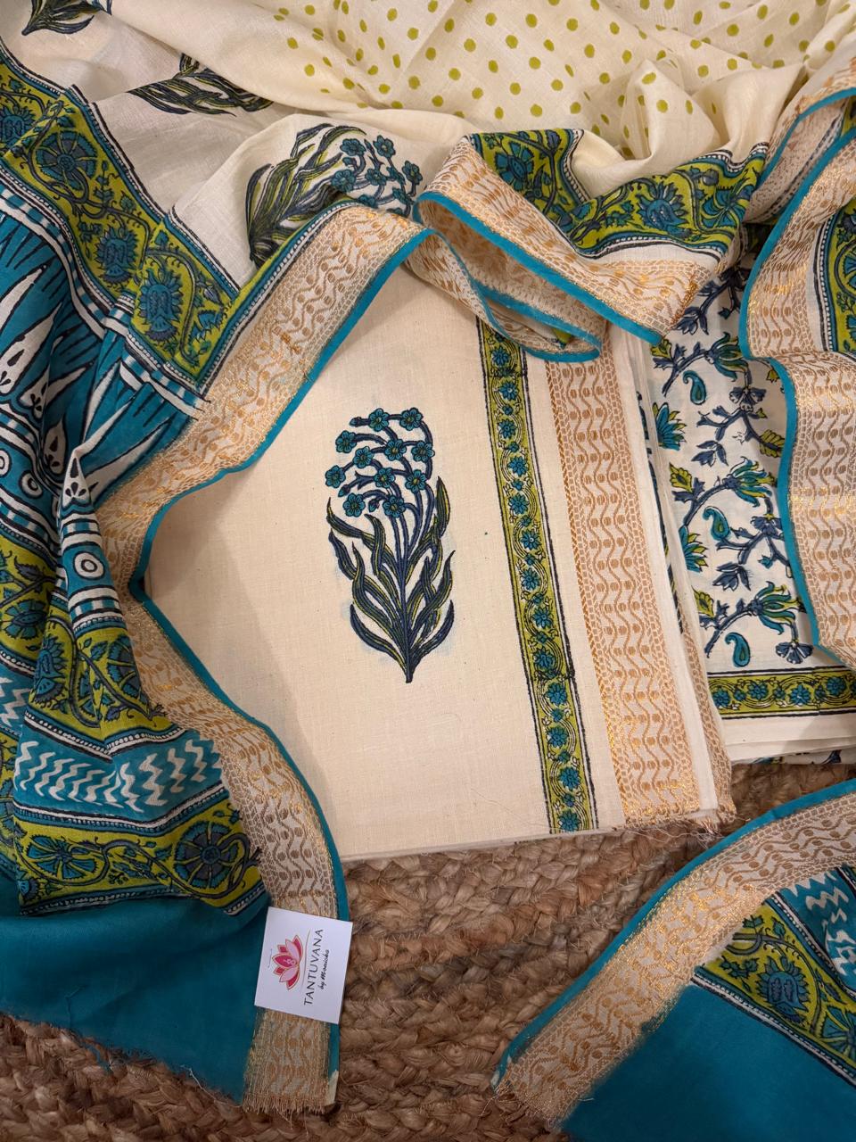 Angled flat lay of cream cotton block print kurta fabric with teal-green floral bouquet motif and golden border with teal piping, alongside teal block print dupatta with green-yellow floral border and cream polka dot fabric 