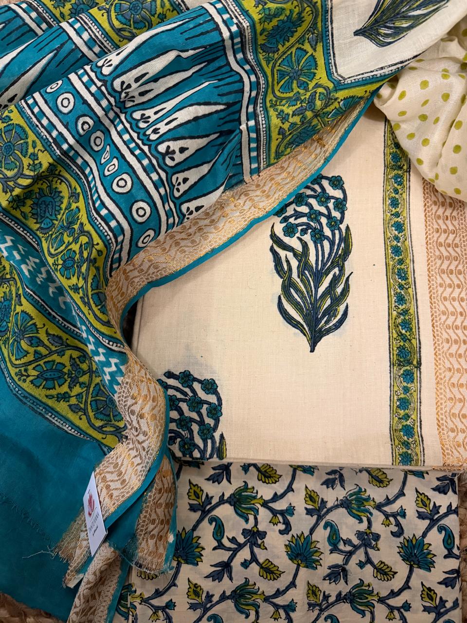 Overhead flat lay of cream cotton block print unstitched suit set with teal block print dupatta draped alongside, showing kurta fabric with teal-green floral bouquet motifs, golden border with teal piping, and cream floral vine bottom fabric 