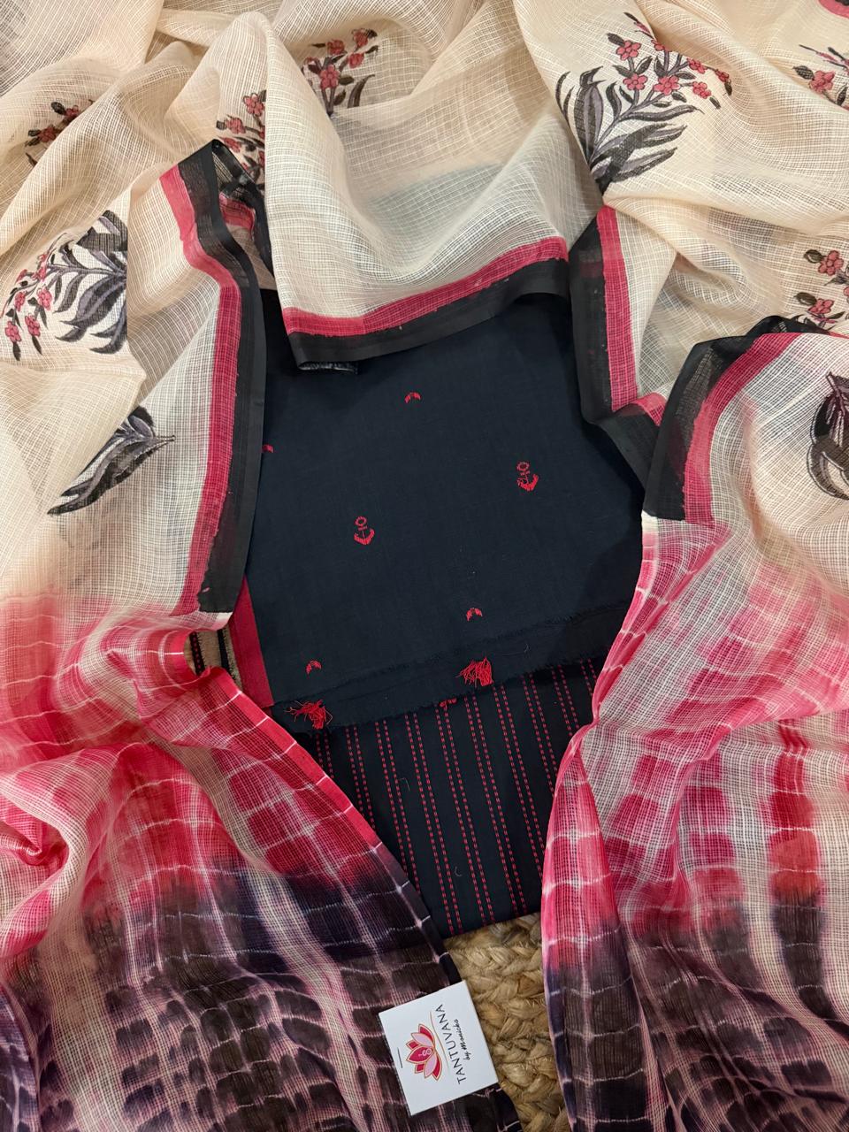 Full flat lay of black and red handloom cotton 3-piece unstitched suit set, Kota Doria dupatta with floral block print, red anchor embroidery kurta, and striped bottom on jute mat