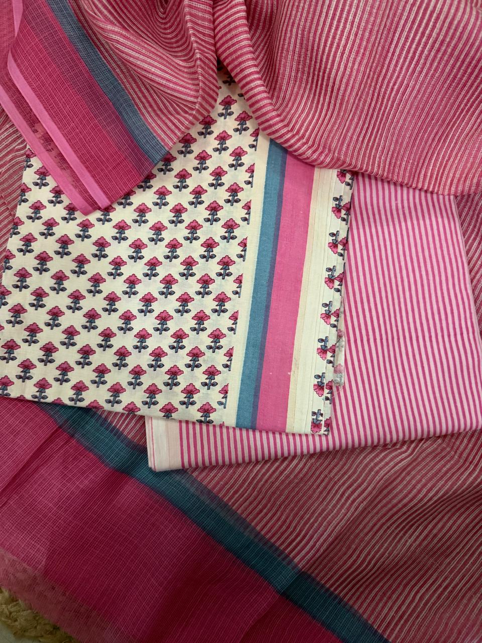 Angled flat lay of cream and pink cotton block print 3-piece unstitched suit set with pink striped Kota Doria dupatta, showing pink floral block print kurta fabric, teal-pink border, and pink striped bottom