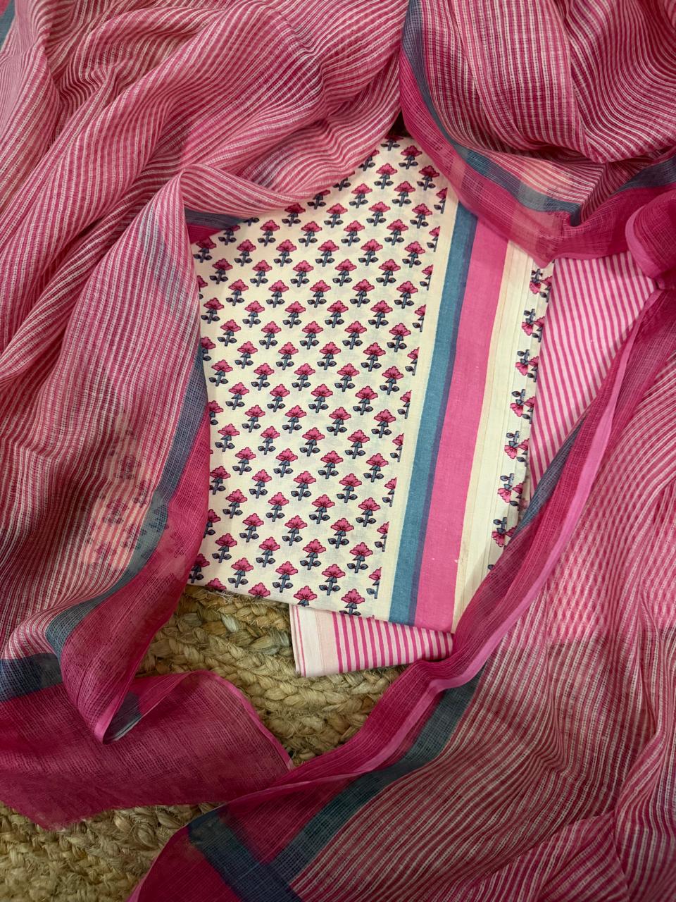 Top-down flat lay of cream and pink cotton block print unstitched suit set with pink striped Kota Doria dupatta framing the set, featuring pink floral block print kurta, teal-pink border panel, and striped bottom