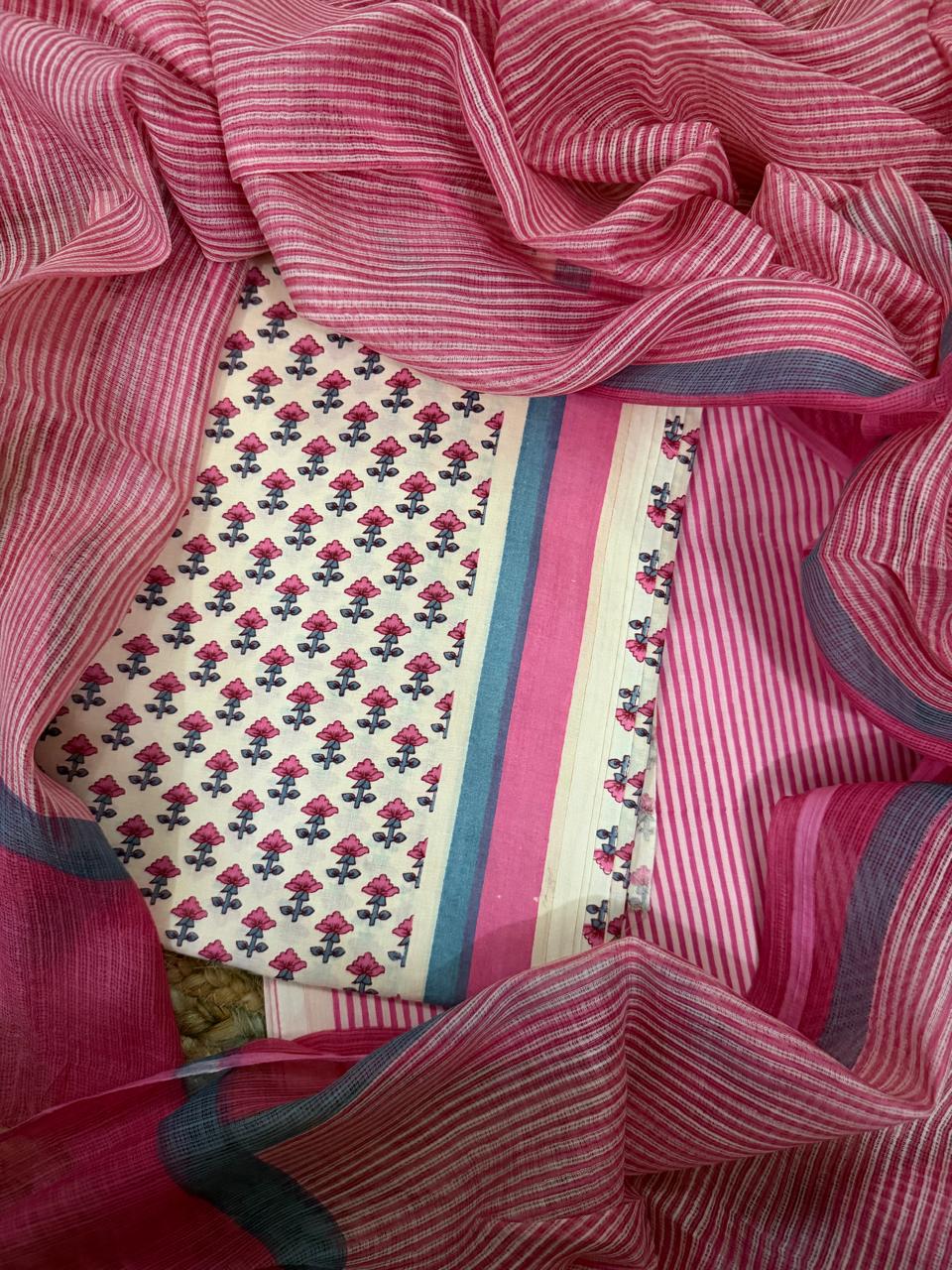 Close overhead flat lay of cream and pink cotton block print unstitched suit set surrounded by pink striped Kota Doria dupatta, highlighting pink floral block print kurta fabric, teal-pink border, and pink striped cotton bottom