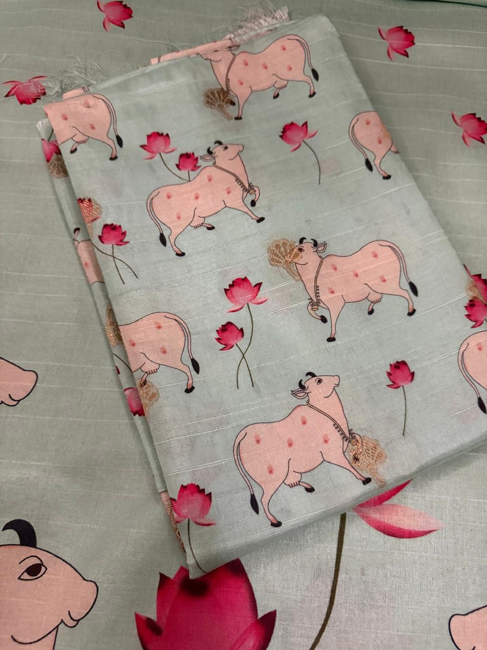 Close flat lay of folded powder green Banarasi semi tussar kurta fabric with Pichwai Kamadhenu cow and pink lotus motifs, placed over the full kurta length showing the allover print