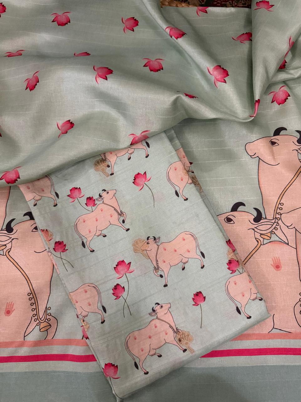 Draped flat lay of powder green Banarasi semi tussar 2-piece unstitched suit showing Pichwai print kurta fabric with pink lotus flowers and decorated Kamadhenu cows, alongside pink-striped border panel