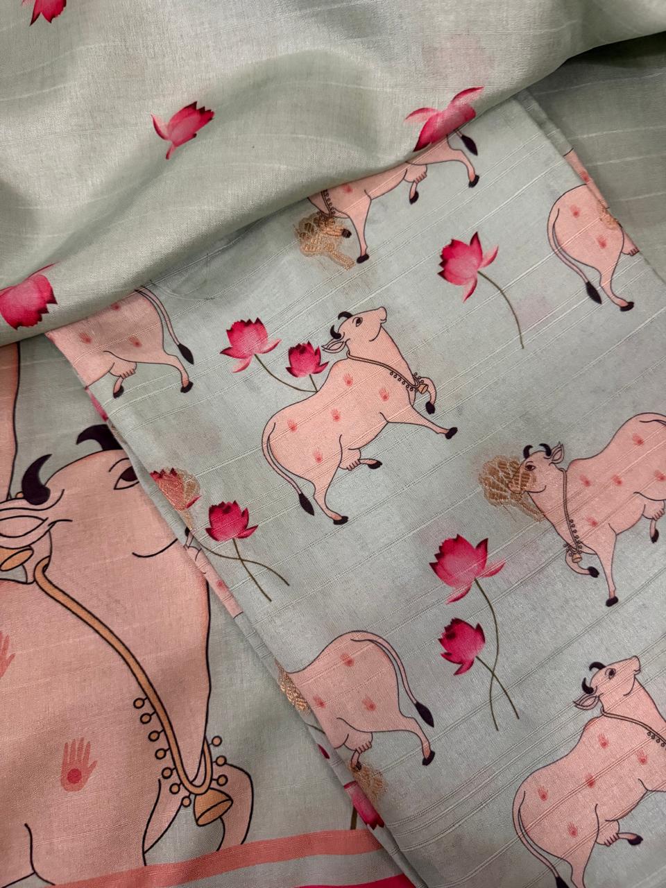 flat lay of powder green Banarasi semi tussar 2-piece unstitched suit with Pichwai print kurta fabric featuring Kamadhenu cows and pink lotus motifs, large Pichwai cow border panel at bottom, and folded bottom fabric on top