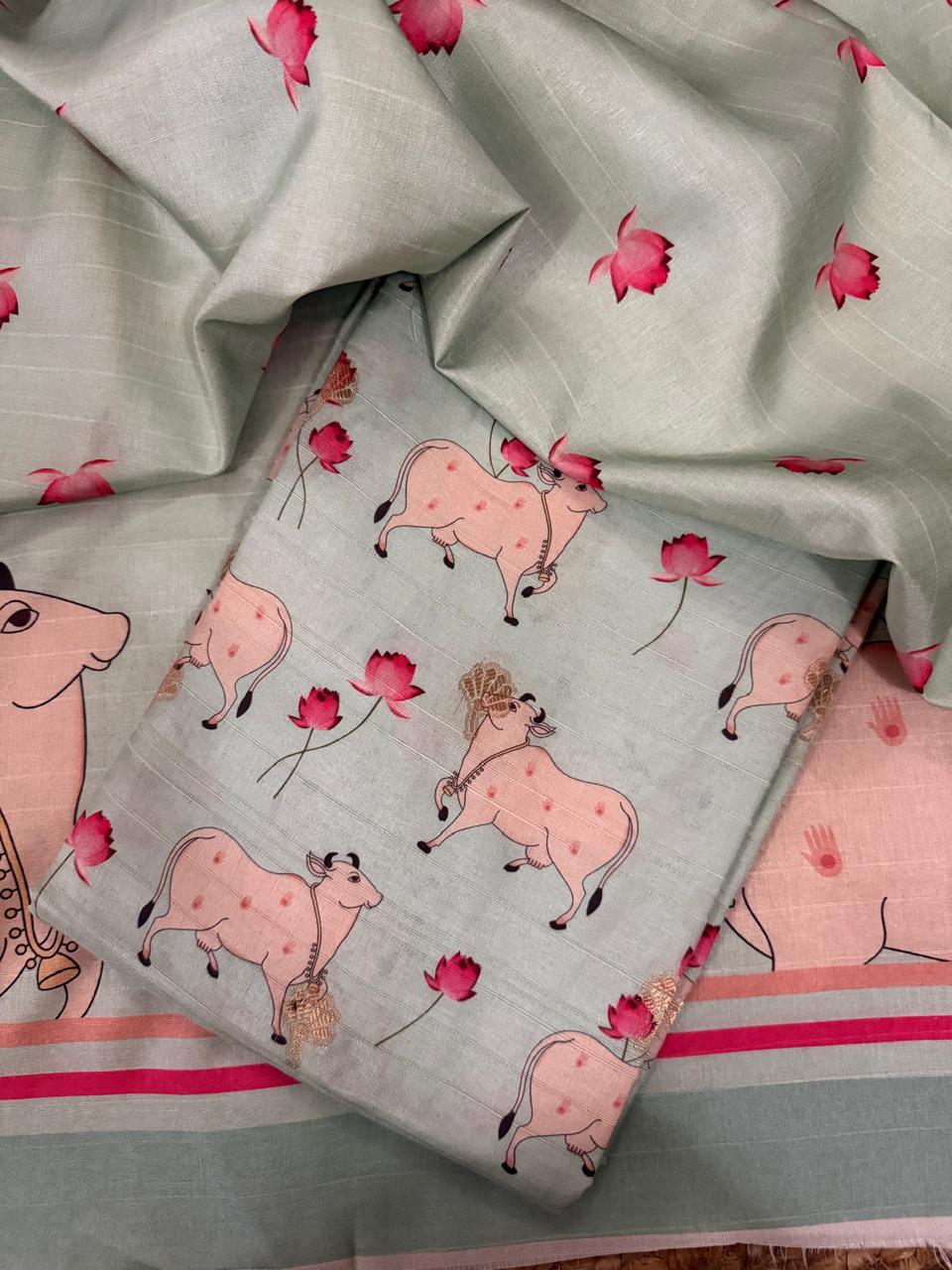 Angled flat lay of powder green Banarasi semi tussar 2-piece unstitched suit with Pichwai print kurta fabric featuring Kamadhenu cows and pink lotus motifs, with pink-striped border panel and large Pichwai cow border visible