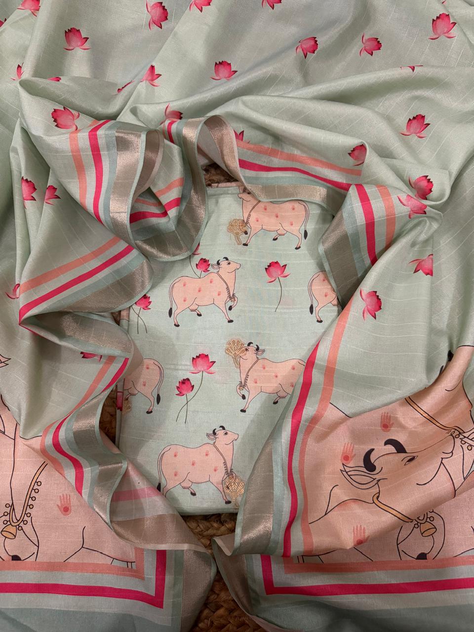 Close-up flat lay of powder green Banarasi semi tussar kurta fabric showing Pichwai Kamadhenu cow motifs with gold tassel embellishments and pink lotus flowers, with large Pichwai cow border panel visible at left edge, highlighting the subtle zari stripe weave texture