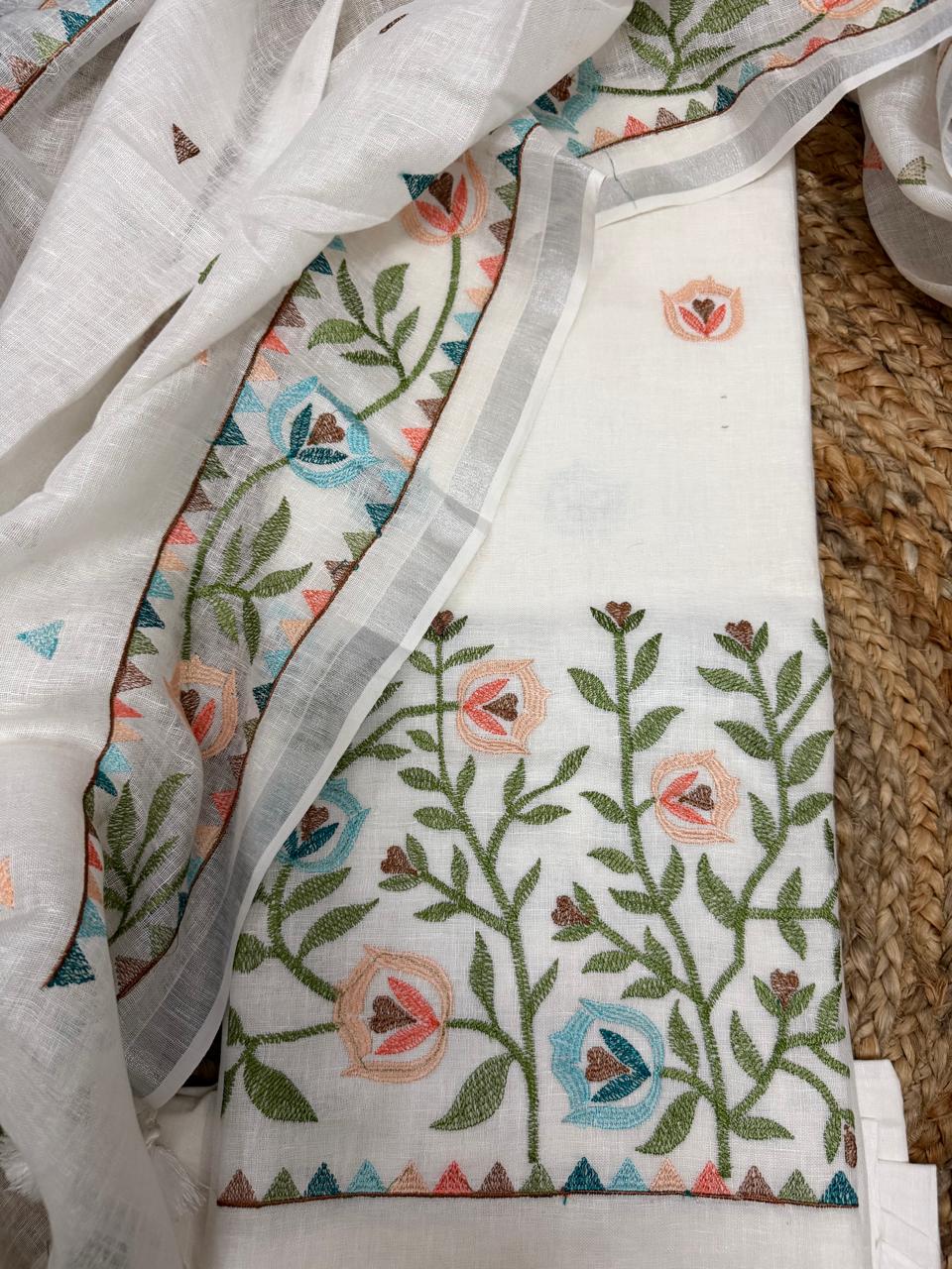 White linen unstitched suit set – angled flat lay on rattan surface showing kurta with multicolour floral vine embroidery hem border and embroidered dupatta