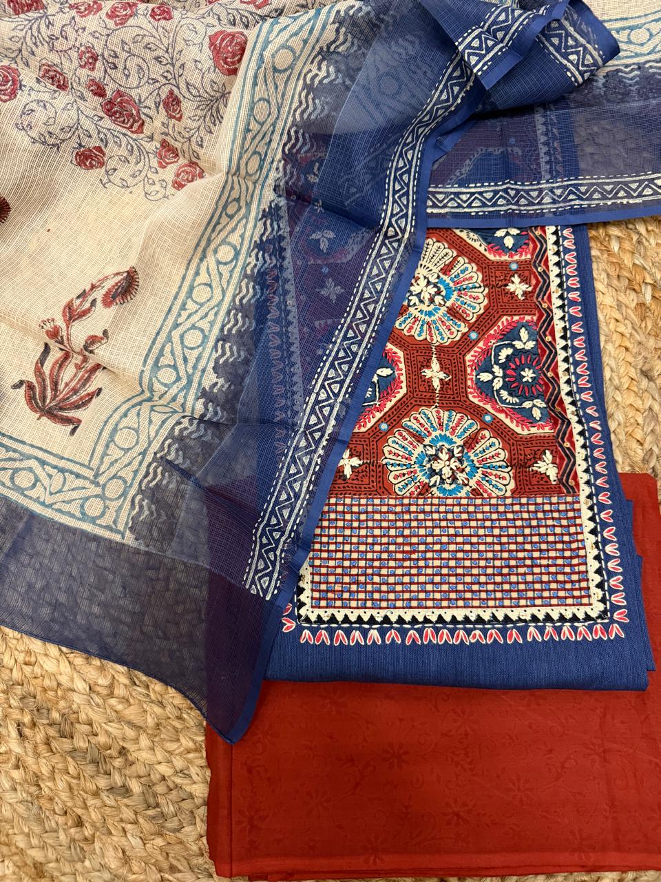 Navy blue and rust 3-piece unstitched suit set – flat lay on rattan showing rust Ajrakh patched neckline panel, navy blue kurta with geometric border, Kota Doria dupatta, and rust salwar fabric